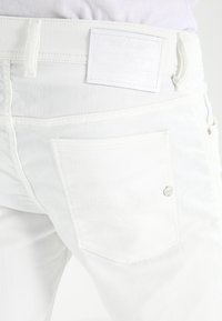 Bílé džíny z denimu mají hladkou texturu, zadní kapsy a bílou koženou nášivku s logem značky. Kovové prvky zdůrazňují design.