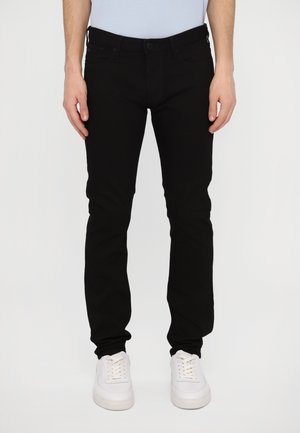Vaqueros de mezclilla negros con un ajuste Slim, cinco bolsillos, pasadores para cinturón y un diseño limpio. Combinados con zapatillas blancas de cordones.