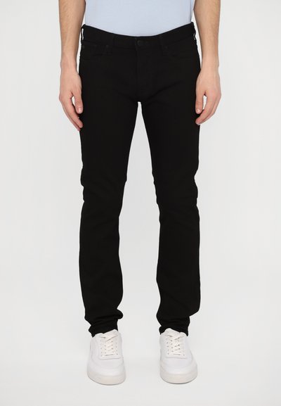 Vaqueros de mezclilla negros con un ajuste Slim, cinco bolsillos, pasadores para cinturón y un diseño limpio. Combinados con zapatillas blancas de cordones.