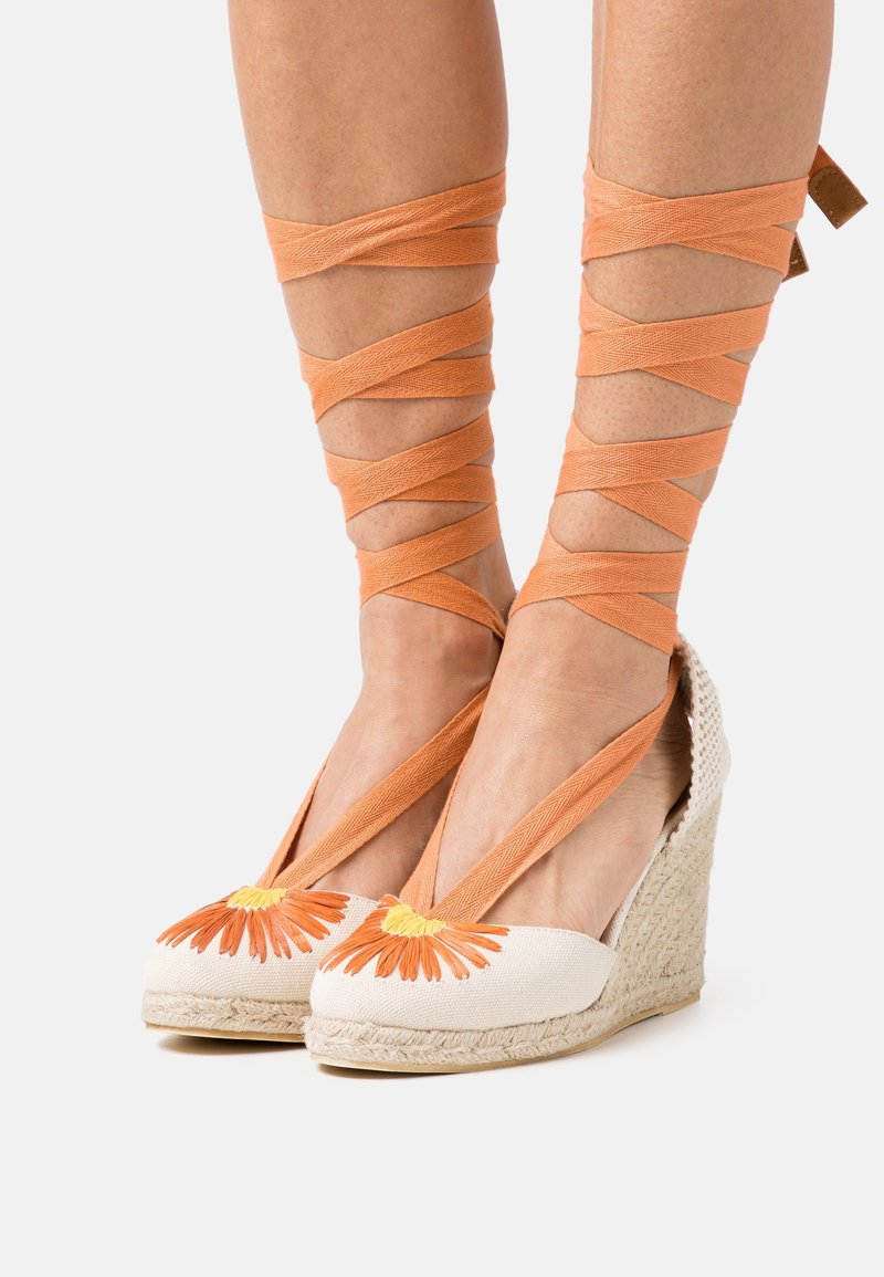 Espadrilles compensées avec broderie de marguerite orange et brides autour de la cheville en tissu orange, portées sur des jambes de peau claire.