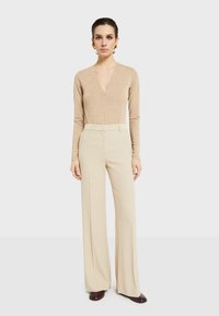 Bež bodi s dugim rukavima i dubokim V-izrezom, uparen s kremastim širokim hlačama visokog struka. Dodaci uključuju zlatne naušnice i tamne cipele.