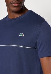 Granatowy t-shirt z krótkim rękawem wykonany z oddychającego materiału, z małym zielonym logo krokodyla oraz poziomym paskiem na wysokości klatki piersiowej.