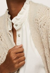 Teksturowany beżowy sweter na guziki, noszony na białej bluzce z falbanianym kołnierzykiem i zatrzaskami, prezentujący warstwowy wygląd i neutralne kolory.