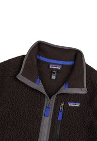 Giacca di pile nera con colletto alto, rifiniture in zip grigie, accenti interni blu e tasca sul petto con zip. Logo Patagonia visibile.