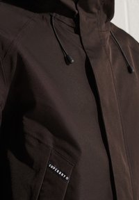 Brązowa kurtka z kapturem wykonana z fakturowanego materiału, z przednią kieszenią z logo "SUPERDRY" oraz regulowanymi sznureczkami przy kapturze.