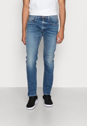 Mann trägt schmal geschnittene hellblaue Jeans, weißes T-Shirt und schwarze Turnschuhe vor einem schlichten weißen Hintergrund, steht aufrecht.
