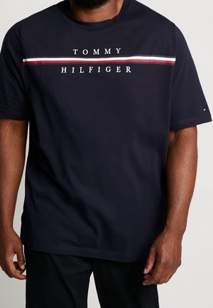 Homme portant un t-shirt Tommy Hilfiger bleu marine avec des rayures blanches et rouges sur la poitrine et le nom de la marque imprimé au-dessus et en dessous des rayures.