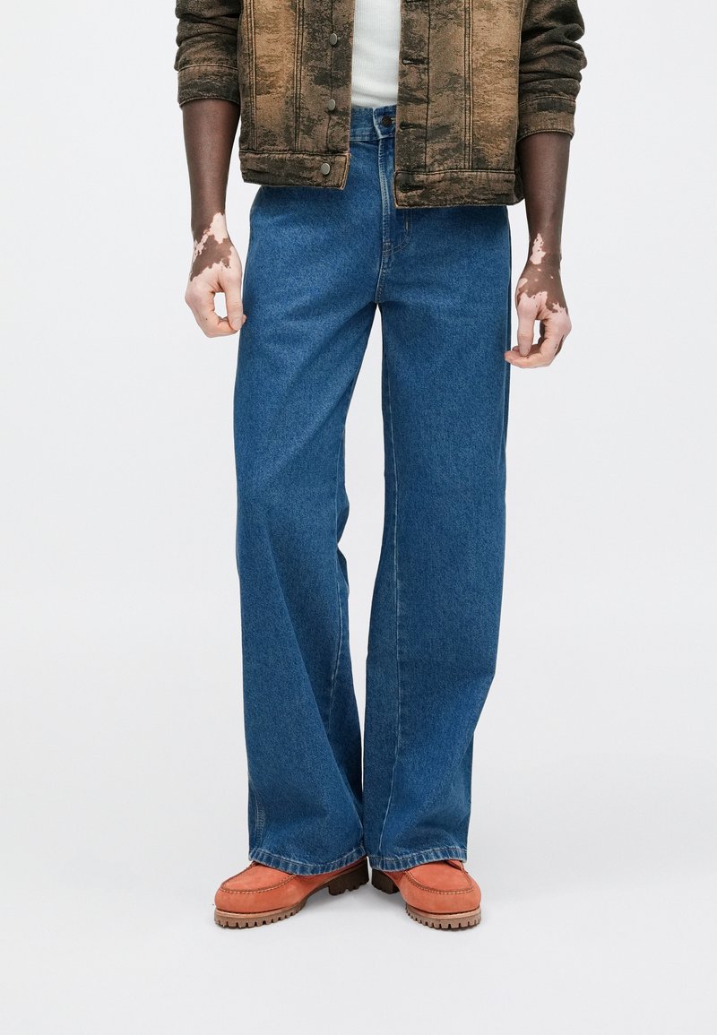 Wide-leg blue jeans made of denim, featuring a classic five-pocket design and a zip fly. The model pairs them with orange shoes.