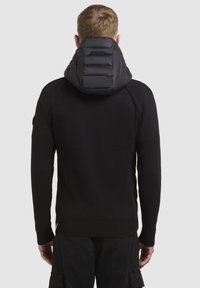 Maglione nero con cappuccio imbottito nero, caratterizzato da sezioni trapuntate, polsini a costine e una silhouette aderente. Il materiale testurizzato fornisce calore.