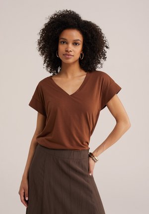 Femme aux cheveux noirs bouclés portant des boucles d'oreilles créoles dorées, un t-shirt marron à col en V et une jupe marron foncé taille haute, posant avec la main sur la hanche.