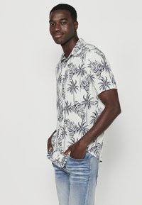 Camicia bianca a maniche corte con stampa di foglie di palma blu scuro, vestibilità ampia, colletto con bottoni, abbinata a jeans azzurri chiari. Tessuto liscio, design casual.