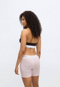 Μαύρο μπουστάκι με λευκή ταινία, σετ με ροζ σορτς μποξέρ που έχουν λευκή ζώνη μέσης με το λογότυπο Calvin Klein. Φαίνεται μπούκλα μαλλιά.