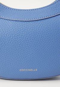 Sac Coccinelle en cuir texturé bleu, présentant une forme courbée et un accent logo doré sur le bord inférieur.
