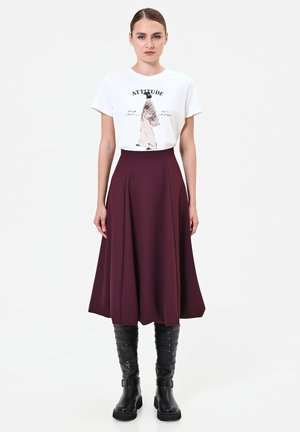 Donna con t-shirt bianca con grafica, gonna midi a linea A viola scuro e stivali neri alti fino al ginocchio su sfondo bianco neutro.
