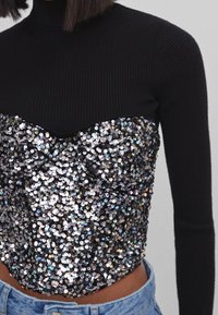 Svart ribbad polotröja med en paljettprydd kroppsektion som har ett skimrande mönster i silver och iriserande accenter. Åtsittande design, kort längd.