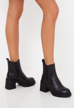 Bottines en cuir noir avec talons blocs portées sur des jambes nues, sur un fond clair uni.