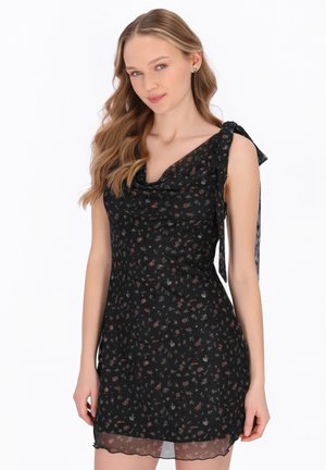 Ung kvinde med bølget blondt hår iført en ærmeløs sort blomsterkjole med bindebånd på skulderen, poserer mod en hvid baggrund.