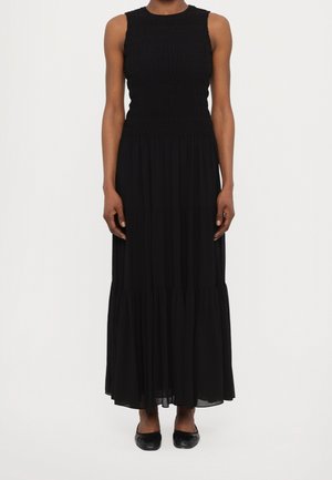 Czarna sukienka maxi bez rękawów z marszczonym gorsetem i wielowarstwową spódnicą, noszona z czarnymi płaskimi butami na osobie stojącej na jednolitym tle.