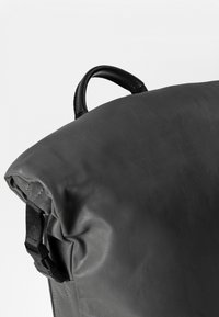 Zaino in pelle nera con una superficie liscia, una patta superiore arrotondata e una chiusura con fibbia laterale. Presenta una maniglia superiore per il trasporto.
