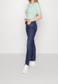 Světlý zelený zkrácený svetr s krátkými rukávy, doplněný tmavě modrými úzkými džínami a bílými teniskami. Hladká textura po celém povrchu.