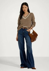 Mulher vestindo um suéter V em ponto cabos castanho, jeans azul de perna larga, cinto castanho, mala de franjas castanha e sapatos castanhos, de pé contra um fundo simples.