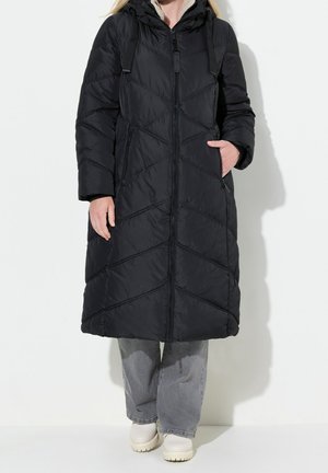 Person iført en lang sort quiltet vinterjakke med hætte, grå bukser og lyse, kraftige støvler, stående mod en hvid baggrund.