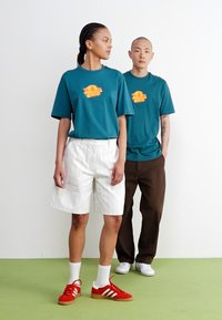 Teal graphic t-shirts with orange sun design, white pinstriped shorts, brown pants, red retro sneakers with white stripes, and green floor.