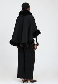 Persona con capelli corti che indossa un mantello nero con bordo di pelliccia, una maglietta bianca a maniche lunghe, pantaloni neri larghi e scarpe con tacco nere, di spalle.