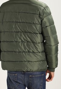 Olivgrön puffjacka med kviltad textur, elastiska ärmmuddar och rund halsringning, bärs över blå jeans. Slät, glänsande tyg.