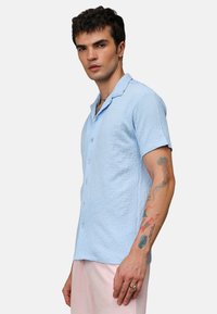 Camisa de manga corta azul claro con tejido texturizado, cierre de botones en la parte delantera y un collar clásico. Presenta mangas enrolladas y un ajuste relajado.
