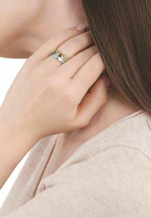 Mano con anillos apilados de oro y plata toca el cabello cerca del cuello, mostrando piel suave y cabello liso castaño.