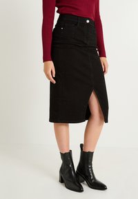 Falda de mezclilla negra con largo hasta la rodilla, abertura frontal y acentos de strass. Combinada con botas negras brillantes hasta el tobillo. Top de canalé burdeos.