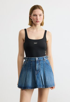 Mlada ženska v črnem brezrokavniku Tommy Jeans in visokim pasom modre denim plise krila, stoječa na belem ozadju.