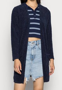 Mörkblå texturerad cardigan, öppen fram, långa ärmar, ihopparad med en randig crop top och en fransad denimkjol med mittlängd vid låren.