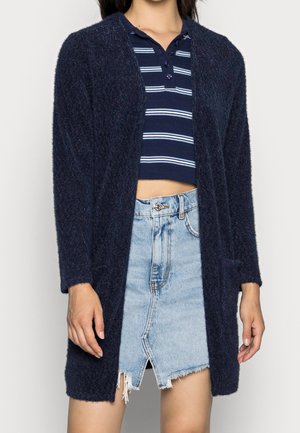 Cardigan texturé bleu marine, à l'avant ouvert, avec des manches longues, associé à un crop top rayé et une jupe en denim effilochée, de longueur mi-cuisse.