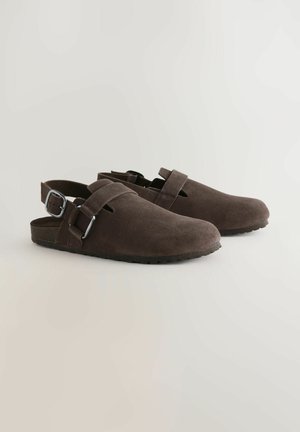 Sabots en daim marron avec sangle réglable, dos ouvert et détails découpés. Semelle plate avec adhérence texturée. Design simple et minimaliste.