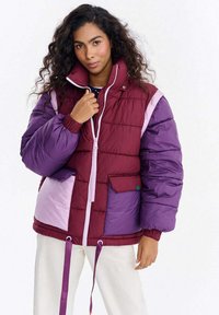 Pikowana kurtka z panelem w odcieniach burgunda, fioletu i lila, zapinana na zamek z przodu, z elastycznymi mankietami i regulacją za pomocą sznureczka u dołu.