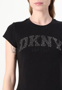 Černé tričko s krátkým rukávem a kulatým výstřihem, se znakem "DKNY NEW YORK" z texturovaných rhinestones a jemného bavlněného materiálu.