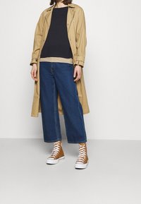 Osoba ubrana w beżowy trencz, warstwowe czarne i w paski bluzki, niebieskie dżinsy z szerokimi nogawkami oraz brązowe wysokie sneakersy, stojąca na tle białym.