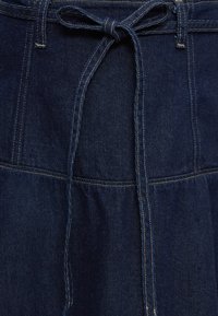 Jupe en denim bleu foncé avec des coutures verticales et une ceinture en tissu nouée à la taille, avec des détails de surpiqûres contrastantes.