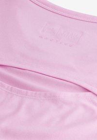Haut de sport rose clair en tissu doux, avec un col rond, une texture subtile et un logo en relief près du col.