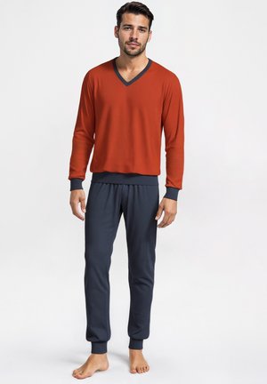 Mann, der barfuß steht und ein rostrotfarbenes langärmliges V-Ausschnitt-Oberteil mit marineblauen Bündchen sowie passende marineblaue Hosen mit Knöchellängen trägt.