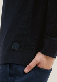 Marineblauwe gebreide trui met een gemêleerd patroon, voorzien van een klein zwart logo-label aan de onderrand en geribbelde manchetten. Hand in een zak geplaatst.