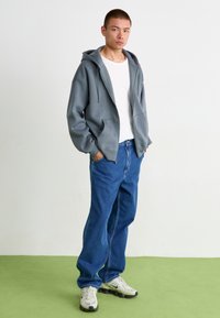 Gray zip-up hoodie, oversized fit, cotton material; paired with blue denim jeans, relaxed style, and white t-shirt underneath.