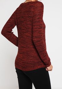 Pull en mailles bordeaux à manches longues avec un motif texturé, silhouette ajustée et un décolleté légèrement arrondi, assorti à un pantalon noir.