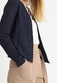 Námořnický texturovaný kardigan s pleteným vzorem, mírně otevřeným předním dílem a dlouhými rukávy, nošený přes světle zbarvenou blůzu a béžové kalhoty s vysokým pasem.