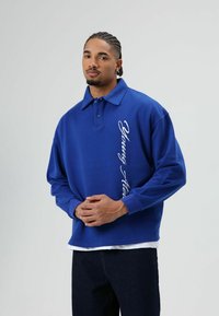 Blauwe katoenen pullover met een kraag, met witte cursieve tekst op de voorkant. Lange mouwen en een losse pasvorm. Donkere spijkerbroek eronder.