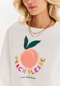 Bílá mikina se zlato-růžovým texturovaným grafikou broskve s zelenými listy, obklopená barevným textem "PEACH PLEASE."