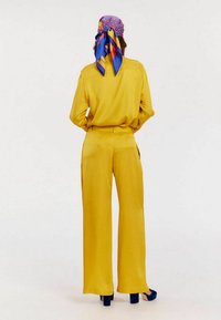 Chemise en polyester jaune et pantalons évasés affichés de derrière, accompagnés d'un foulard à motifs bleu et jaune. Texture lisse, coupe décontractée.