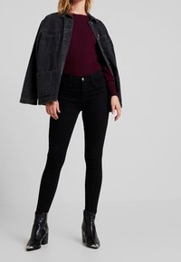 Schwarze Jeansjacke mit zwei aufgesetzten Taschen, bordeauxrotes, geripptes Langarmoberteil, schwarze Skinny-Jeans und schwarze Knöchelstiefel mit metallischen Akzenten.